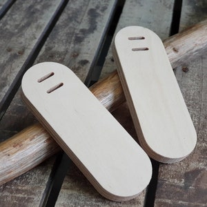 May include: Two unfinished wooden oval-shaped pieces with two holes each. The pieces are lying on a wooden surface.