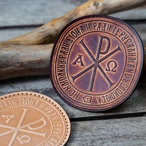 Op de afbeelding: Twee ronde leren onderzetters in bruin en beige tinten. De onderzetters hebben een christelijk symbool met de tekst "HS  ЧCOKOYHCKAЯ  H  PAZH  CHONA  H  PAZH  IEPCYCAZH" rond de rand.