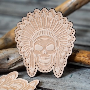 Op de afbeelding: Een leren patch met een gedetailleerd reliëf van een schedel die een hoofdtooi met veren draagt. De patch is beige en heeft een gestikte rand.