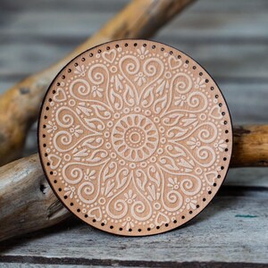 May include: A round wooden coaster with a white mandala design. The coaster has a brown border and a light brown background.