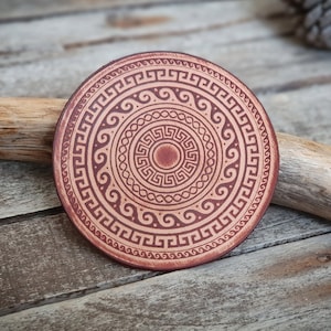 Peut inclure: Un dessous de verre rond en cuir avec un motif géométrique dans des tons de brun. Le motif présente des cercles concentriques de motifs répétitifs, y compris un motif de clé grecque.