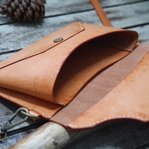 May include: A light brown leather envelope-style bag with a flap closure and a snap button. The bag has a white stitching detail and a small strap with a metal clasp.
