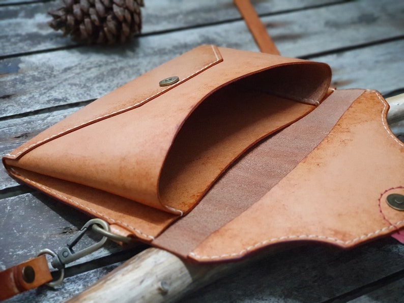 May include: A light brown leather envelope-style bag with a flap closure and a snap button. The bag has a white stitching detail and a small strap with a metal clasp.