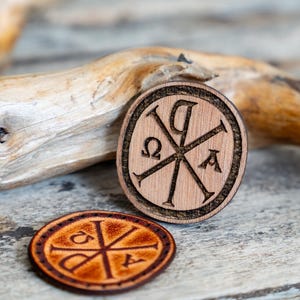 May include: Two circular tokens featuring the Chi Rho symbol with Alpha and Omega letters. One token is made of light-colored wood with a dark laser-engraved design, while the other is made of dark brown leather with a darker embossed Chi Rho emblem.
