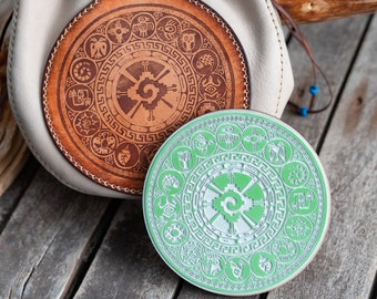 May include: Two round, decorative coasters with a green and brown design. The coasters feature a circular pattern with a central symbol and a border of smaller symbols. The coasters are made of leather and wood.