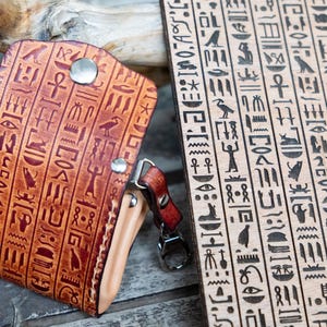 May include: A brown leather key holder with a metal snap closure and a key ring. The key holder is embossed with hieroglyphs. Next to it is a wooden plank with more hieroglyphs carved into it.