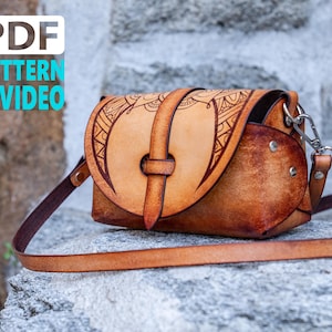 Puede incluir: Un bolso de cuero marrón cruzado con un diseño decorativo en la solapa delantera. El bolso tiene una correa y un cierre de metal. El bolso está sobre una superficie de piedra.