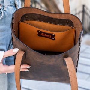 May include: A brown leather tote bag with a light brown interior pocket. The bag has two handles and a stitched design along the sides. The interior pocket has a brown label with the word "Eleana".