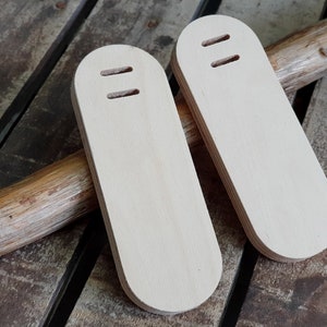 May include: Two wooden, light brown, oval-shaped, blank, unfinished,  wood blanks with two holes each. The blanks are sitting on a wooden surface.