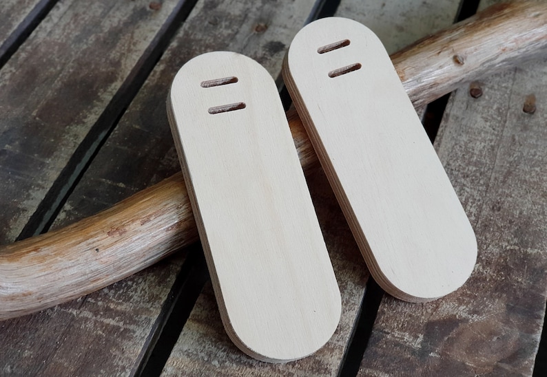 May include: Two wooden, light brown, oval-shaped, blank, unfinished,  wood blanks with two holes each. The blanks are sitting on a wooden surface.