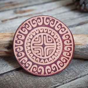 May include: A round, brown leather coaster with a decorative, intricate design in a lighter brown color. The design features a central cross shape surrounded by concentric circles of swirling patterns.