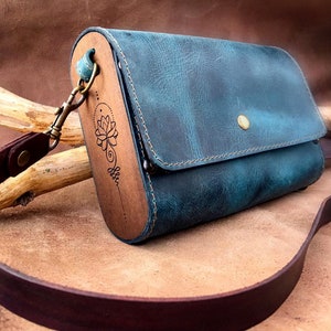 May include: A teal blue leather crossbody bag with a wooden flap featuring a laser-etched lotus flower design. The bag has a brown leather strap and a brass clasp.