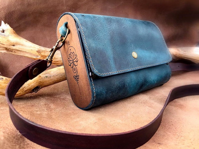 May include: A teal blue leather crossbody bag with a wooden flap featuring a laser-etched lotus flower design. The bag has a brown leather strap and a brass clasp.