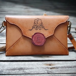 May include: A tan leather envelope-style crossbody bag with a decorative seal and the letter "E" engraved on it. The bag has a long strap and a metal clasp.