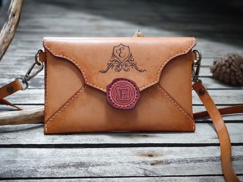May include: A tan leather envelope-style crossbody bag with a decorative seal and the letter "E" engraved on it. The bag has a long strap and a metal clasp.