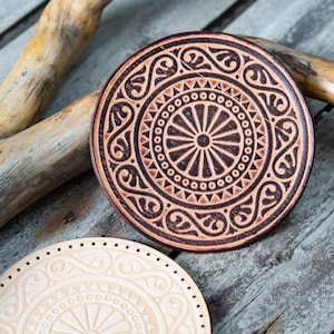 May include: Two round leather coasters with intricate geometric and floral designs. The coaster on the left is light beige with a white design, while the coaster on the right is brown with a black design.