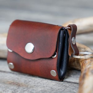 May include: A small, brown leather pouch with a silver key ring attached. The pouch has a curved flap secured by a silver stud and is accented with additional silver rivets. The leather has a rich, reddish-brown hue.