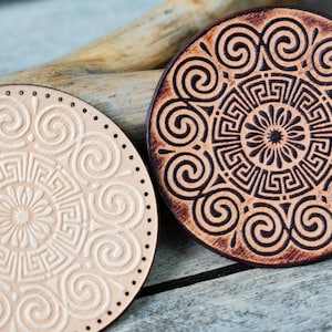 May include: Two round leather coasters with a decorative design of a mandala with a central flower and swirling patterns. One coaster is light brown with a white design, and the other is dark brown with a light brown design.