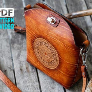 May include: A brown leather crossbody bag with a decorative circular design. The bag has a long, adjustable strap and a silver clasp. The image also includes the text "PDF PATTERN VIDEO". The bag is likely made from high-quality leather.