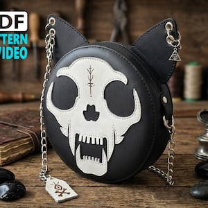Könnte beinhalten: Eine runde schwarze Tasche mit Katzenohren und einer weißen Totenkopf-Grafik auf der Vorderseite. Die Tasche hat einen silbernen Kettenriemen, einen kleinen dreieckigen Anhänger und ein rechteckiges Etikett mit Totenkopf-Symbol. Text PDF PATTERN VIDEO.