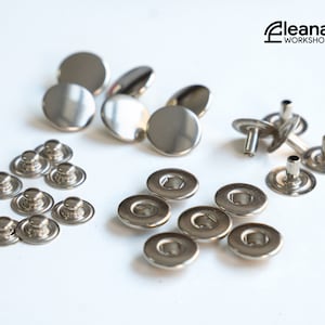 Op de afbeelding: Assortiment van zilveren metalen drukknopen, waaronder ronde drukknopen, tongdrukknopen en pen-drukknopen.