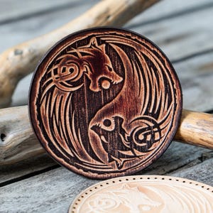 Puede incluir: Un posavasos de madera con un diseño yin yang que presenta dos lobos, uno con alas, en un color marrón oscuro.