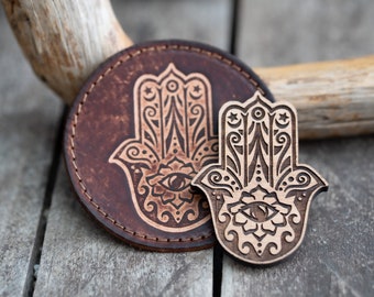 Può includere: Due ciondoli a forma di mano di hamsa, uno è un sottobicchiere in pelle marrone con un design di hamsa nero, l'altro è un ciondolo di hamsa in legno con venature di legno marrone e un design di hamsa nero.