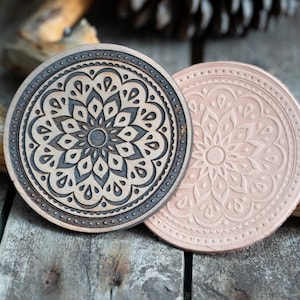 May include: Two round leather coasters with a mandala design. One coaster is brown with a black mandala design, and the other is light brown with a white mandala design.