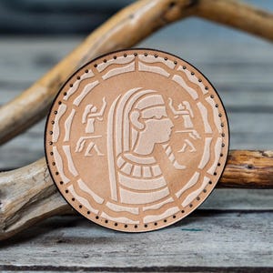 Könnte beinhalten: Ein runder Lederuntersetzer mit einem detaillierten Design einer ägyptischen Frau mit Kopfbedeckung und zwei Figuren auf jeder Seite. Der Untersetzer ist braun mit einem weißen Design.