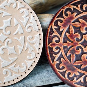 Op de afbeelding: Twee ronde houten onderzetters met ingewikkelde gesneden ontwerpen. De ene onderzetter is lichtbruin met een wit ontwerp, de andere is donkerbruin met een zwart ontwerp.