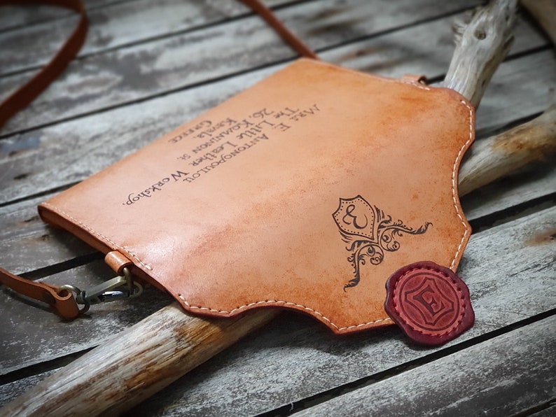 May include: A tan leather crossbody bag with a decorative seal and the text "The Little Leather Workshop. MR. E ANTONOPOULOU 26. KOMNINON S Kamla. GREECE" engraved on the front.