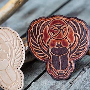 May include: Two leather patches featuring an Egyptian scarab beetle design. The patch on the left is light brown and the patch on the right is dark brown.