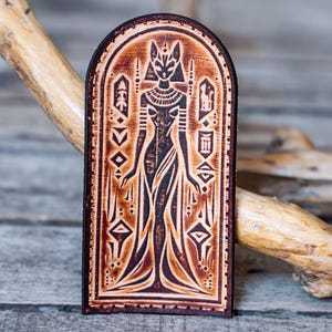May include: A rectangular leather patch with rounded top edges featuring a dark brown embossed illustration of an Egyptian deity figure. The design includes geometric patterns and symbols surrounding the central figure, with visible stitching along the border.