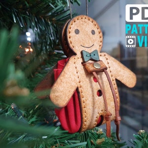 May include: A gingerbread man ornament made of leather with a red felt pocket and brown leather straps. The ornament is decorated with brown stitching and a green bow tie.