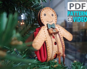 May include: A gingerbread man ornament made of leather with a red felt pocket and brown leather straps. The ornament is decorated with brown stitching and a green bow tie.