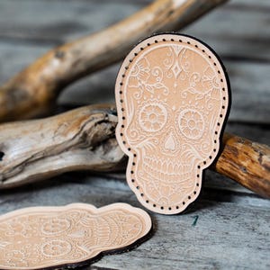 Puede incluir: Dos parches de cuero marrón en forma de calavera de azúcar con intrincados diseños florales blancos. Las calaveras tienen una cara detallada con una nariz en forma de corazón y una sonrisa  dentada.