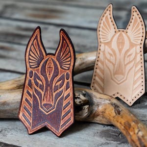 May include: Two wooden Anubis head carvings, one in brown and one in natural wood. Both carvings feature detailed lines and an ankh symbol.