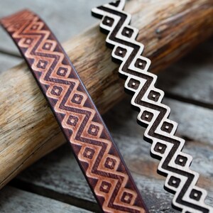 Puede incluir: Primer plano de dos objetos decorativos de madera con patrones geométricos. Uno es una correa de cuero marrón con un diseño de zigzag y diamantes que se repite. El otro es una pieza de madera en forma de zigzag con un patrón similar en marrón oscuro.