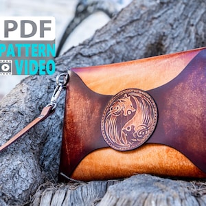 May include: A handcrafted leather crossbody bag with a unique design. The bag features a circular emblem with an engraved animal motif. The bag has a long, adjustable strap and a smaller wrist strap. The bag is made of brown and tan leather.