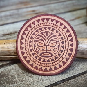 Può includere: Un sottobicchiere rotondo in pelle con un design marrone e beige. Il design presenta un viso stilizzato con motivi geometrici intorno.