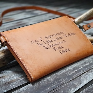 May include: A tan leather crossbody bag with a strap and a brass clasp. The bag has the text "Mrs. E. Antonopoulou The Little Leather Workshop, 26, Komninon St, Kavala, GREECE" engraved on the front.