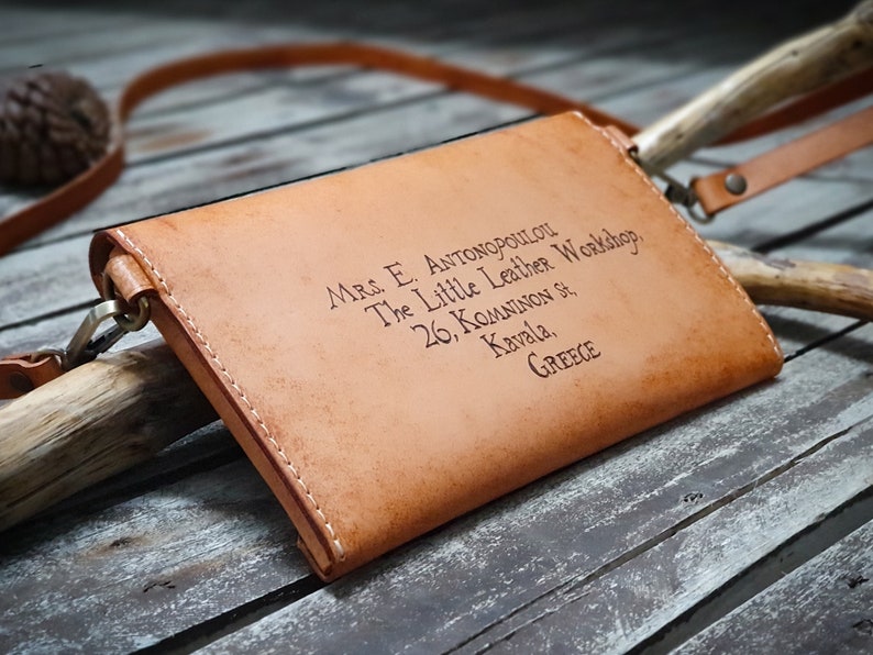 May include: A tan leather crossbody bag with a strap and a brass clasp. The bag has the text "Mrs. E. Antonopoulou The Little Leather Workshop, 26, Komninon St, Kavala, GREECE" engraved on the front.