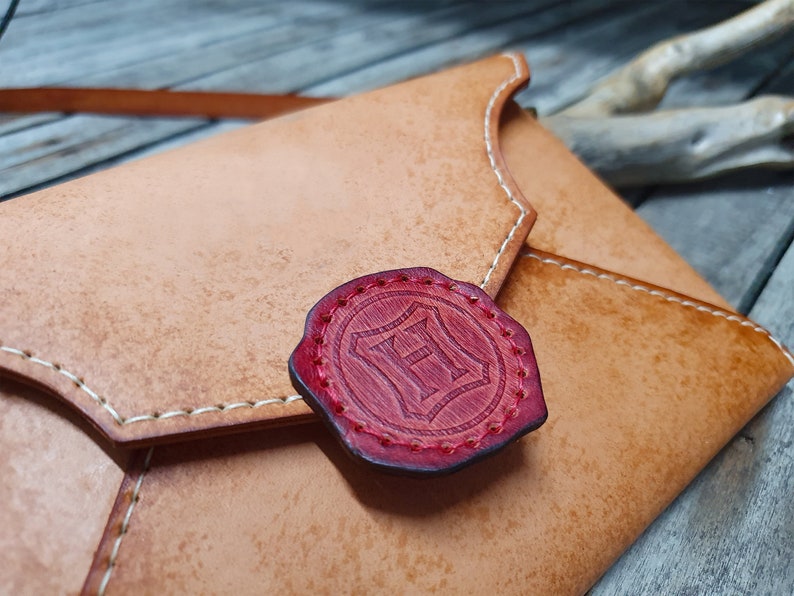 May include: A tan leather envelope with a red wax seal featuring the letter "H" in a shield design.