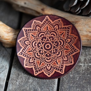 Peut inclure: Un dessous de verre rond en cuir marron foncé avec un motif mandala détaillé de couleur marron clair. Le dessous de verre est posé sur une surface en bois vieilli, mettant en valeur le motif floral complexe.