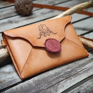 May include: A tan leather envelope-style crossbody bag with a decorative seal and a monogrammed design. The bag has a strap and is made of genuine leather.