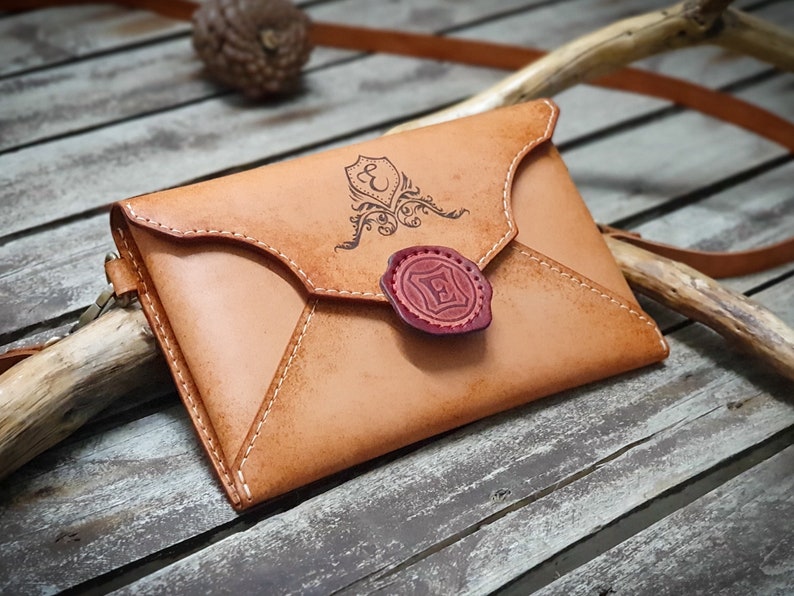 May include: A tan leather envelope-style crossbody bag with a decorative seal and a monogrammed design. The bag has a strap and is made of genuine leather.
