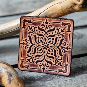 May include: Square, brown leather coaster with a detailed, symmetrical floral design. The coaster has a dark border and a lighter, embossed pattern in the center. The design is reminiscent of a mandala or tile.