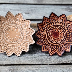 Puede incluir: Dos posavasos de madera con forma de flor. Uno es de color beige claro con un diseño en relieve detallado. El otro es de color marrón oscuro con un diseño en relieve similar, pero más oscuro. Ambos están sobre una superficie de madera envejecida.