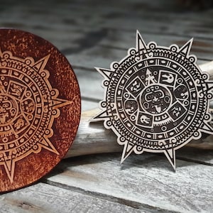 Puede incluir: Dos placas redondas decorativas de madera con diseños intrincados. Las placas presentan un patrón circular con una figura central y símbolos circundantes. Una placa es marrón con un tono rojizo, la otra es de un marrón más claro.