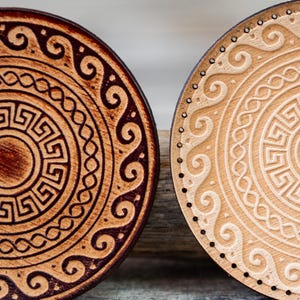 Puede incluir: Dos posavasos redondos de madera con un diseño geométrico. Un posavasos es marrón con un diseño marrón oscuro, y el otro es marrón claro con un diseño blanco.